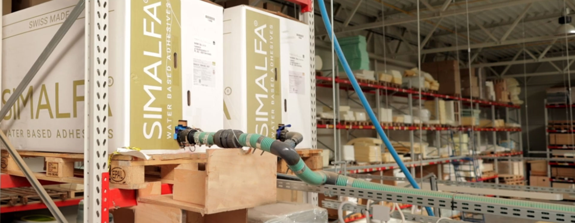 Produktionshalle mit großen 500kg Container von SIMALFA® wasserbasierten Klebstoffen, die an eine Produktionsanlage angeschlossen sind.