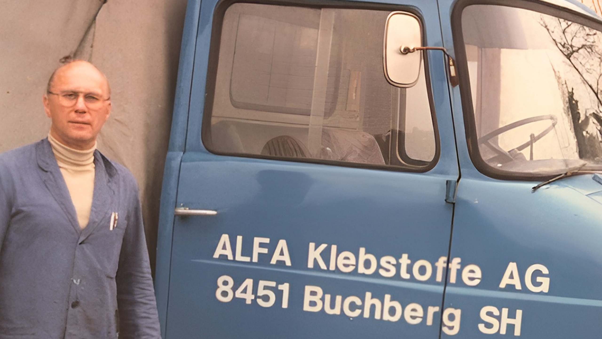 Historic photo of an employee in work clothes standing in front of an ALFA vehicle, taken in front of the company headquarters in Buchberg.