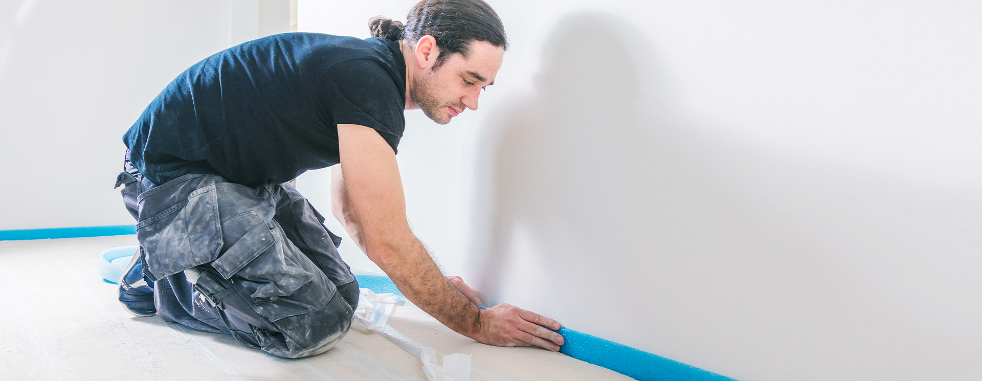 A man kneels on a floor and installs a blue foam strip along a wall.